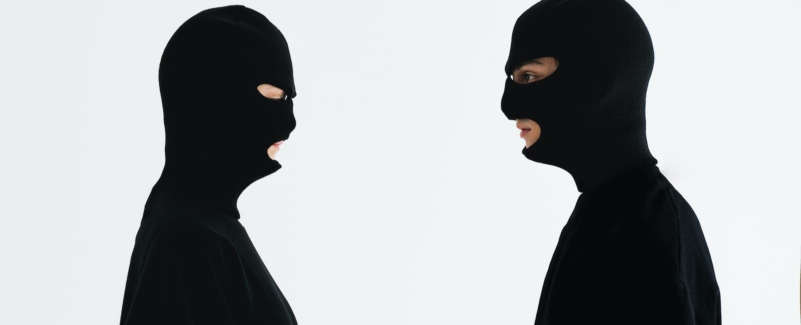 Two people in black balaclavas facing each other against a plain white background