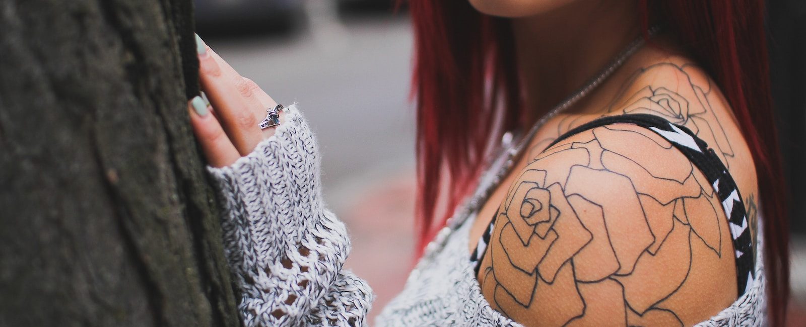 Woman with red hair wearing knitted sweater, showing floral shoulder tattoo and ring, standing by a tree on city street