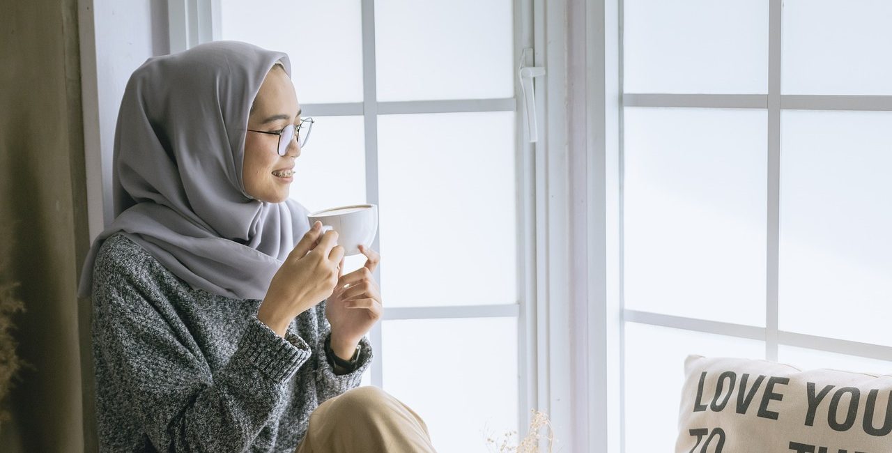 Woman in grey hijab sitting by window, holding a coffee mug, enjoying a peaceful moment indoors without full stop