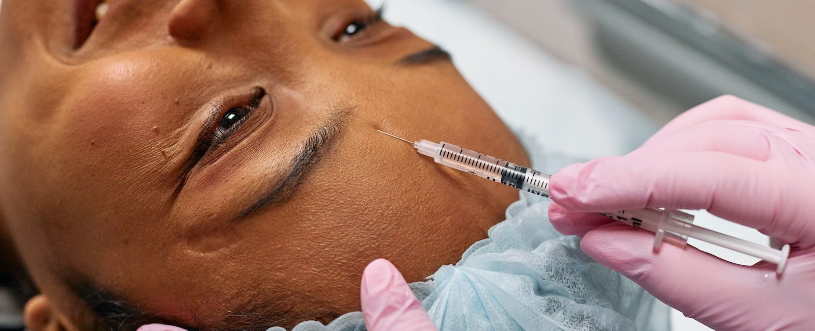 Person receiving a cosmetic injection on the forehead in a clinic setting, wearing protective cap and gloves