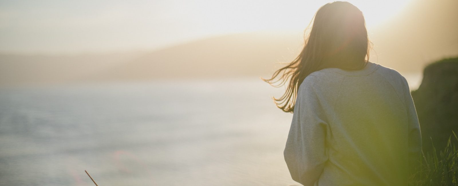 Woman sitting on grass overlooking serene ocean at sunset with sunlight illuminating the scene
