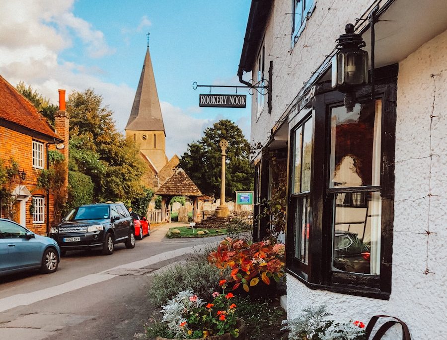Quaint English village street with church spire, historic buildings, and floral displays in wooden barrels