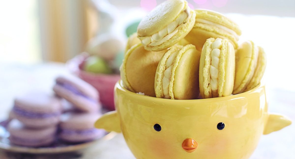 Yellow chick-shaped bowl filled with yellow macarons on floral tablecloth, purple macarons in background