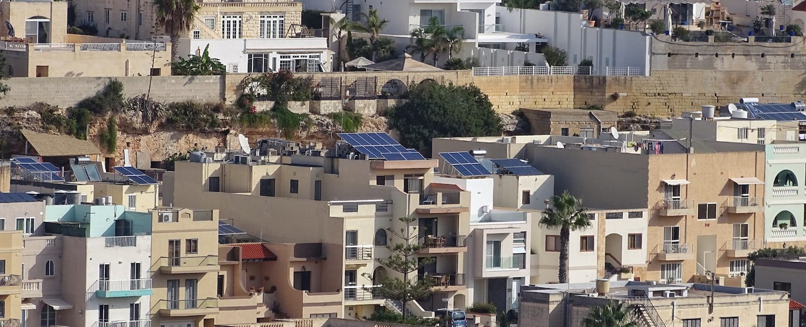 Hillside Mediterranean village with solar panels on white and beige buildings surrounded by greenery and palm trees