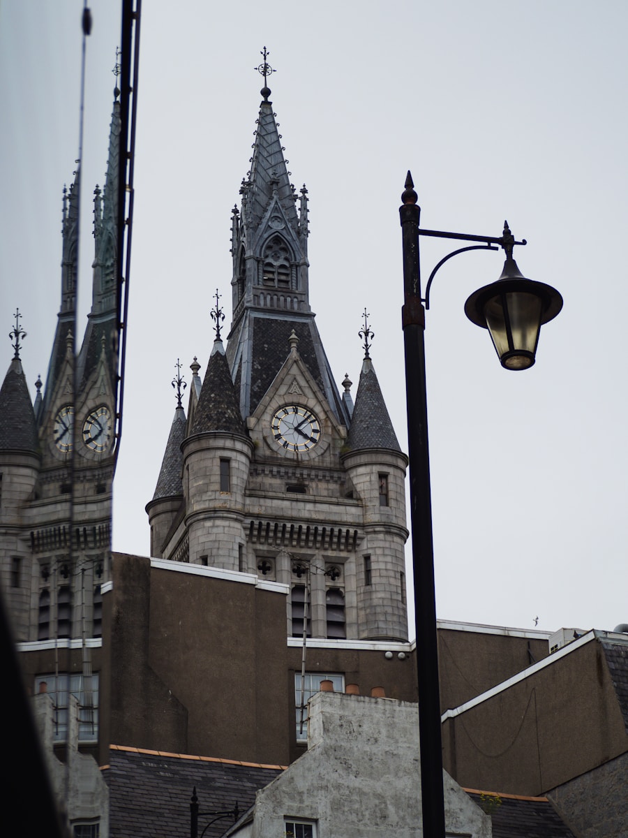 Gothic clock tower with ornate spires against cloudy sky, reflected in glass alongside street lamp