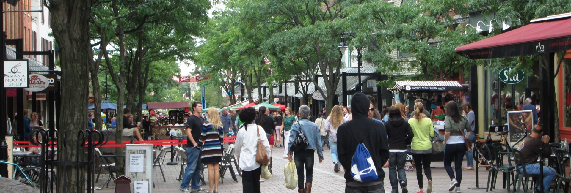 People walking on a bustling pedestrian street lined with shops, trees, and outdoor seating