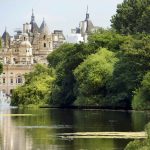 Historic building with spires behind lush park trees overlooking a serene pond with a water fountain