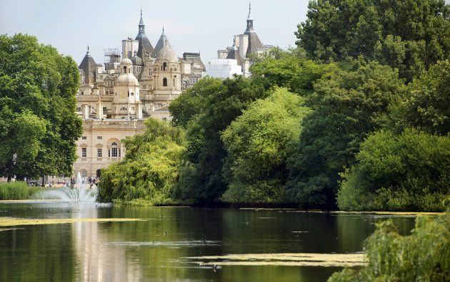 Historic building with spires behind lush park trees overlooking a serene pond with a water fountain