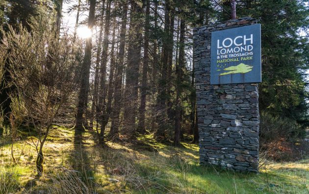 Signage of Loch Lomond and The Trossachs National Park with sun shining through forest trees