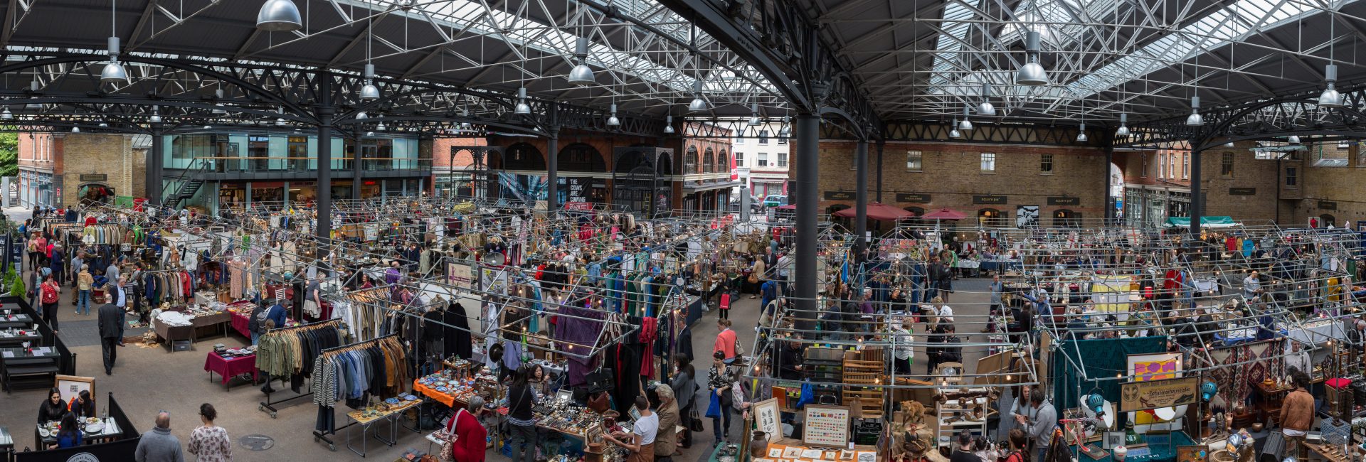 Bustling indoor market with diverse clothing, antiques, and people shopping under a metal roof in an urban setting