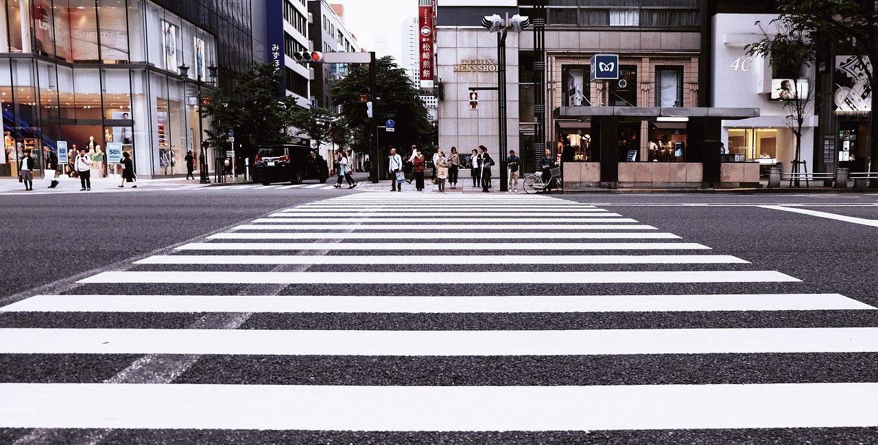 Wide zebra crossing on a busy city street with people waiting to cross, surrounded by modern buildings and shops