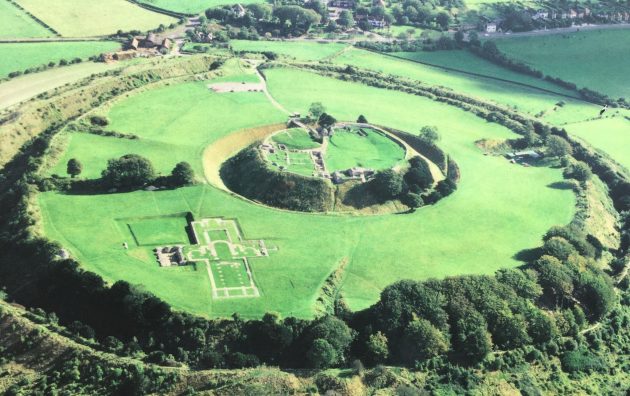 Aerial view of historic English earthwork and ruins amidst lush green landscape near village houses