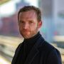 Man in black coat and turtleneck in urban setting, gazing towards camera, blurred city background