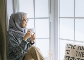 Woman in grey hijab sitting by window, holding a coffee mug, enjoying a peaceful moment indoors without full stop