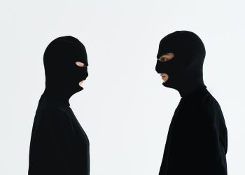 Two people in black balaclavas facing each other against a plain white background