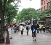 People walking on a bustling pedestrian street lined with shops, trees, and outdoor seating