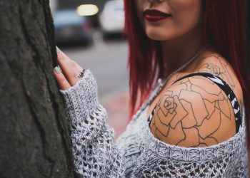 Woman with red hair wearing knitted sweater, showing floral shoulder tattoo and ring, standing by a tree on city street