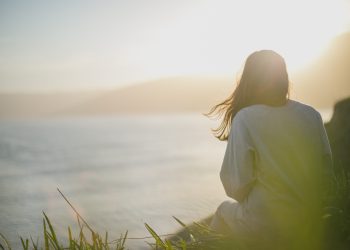 Woman sitting on grass overlooking serene ocean at sunset with sunlight illuminating the scene