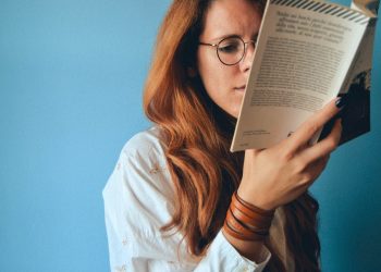 Woman with long hair and glasses reading a book against a blue wall background