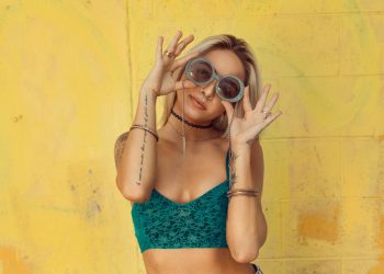 Stylish woman in green top with tattoos playfully holding round sunglasses against a vibrant yellow wall