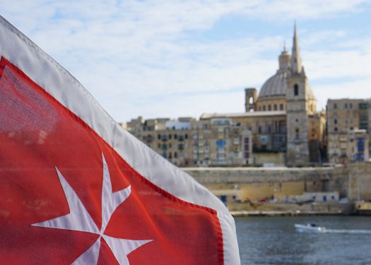 Maltese flag with Valletta skyline in background displaying historic architecture under a blue sky